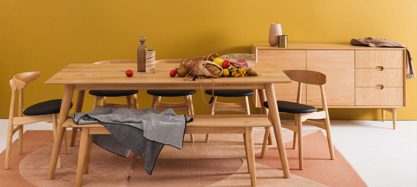Modern wooden dining set with chairs and bench in front of mustard yellow wall, featuring decorative items and fresh bread on the table.