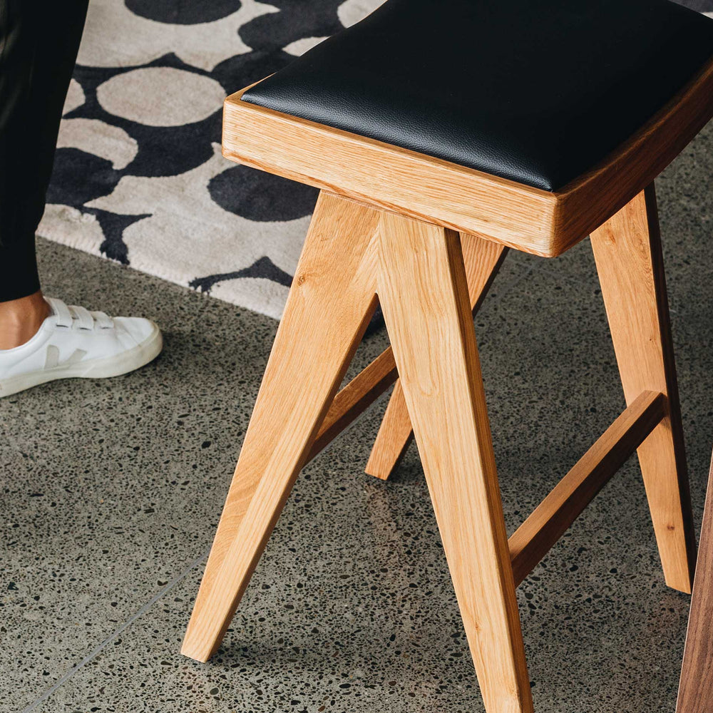 Wooden stool with black seat next to a person walking on a concrete floor.