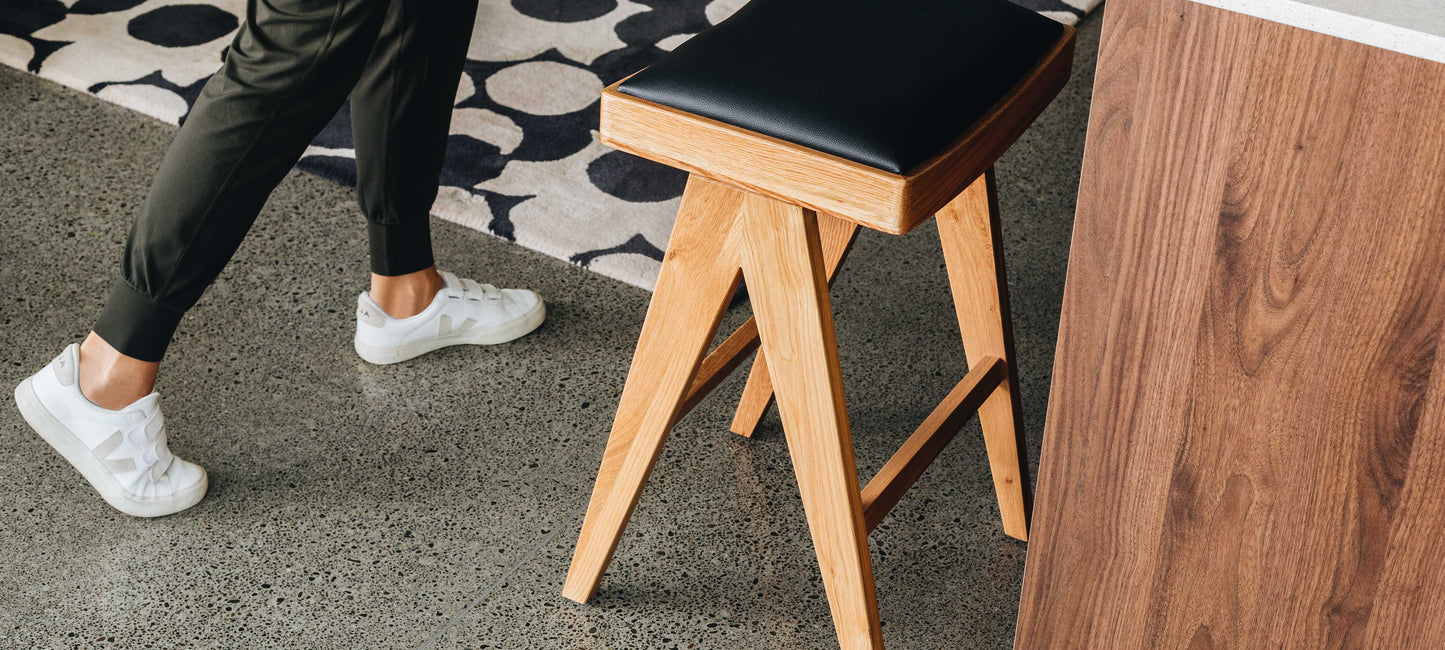 Wooden stool with black seat next to a person walking on a concrete floor.