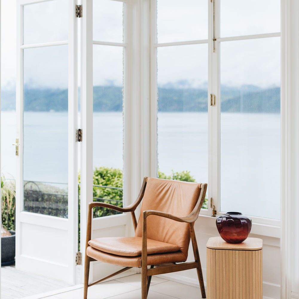 
                      
                        Finn Armchair in a sunlit room by large windows with scenic lake view, mid-century design in ash wood and vintage leather.
                      
                    