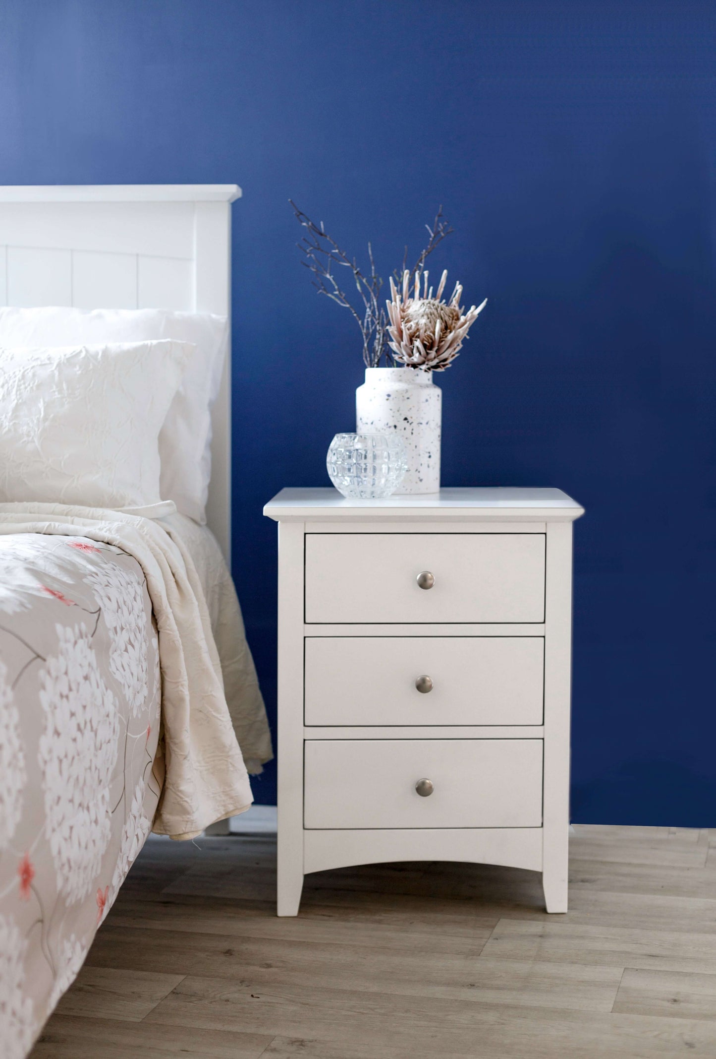 White bedside table with three drawers and decorative vase against a blue wall in a cozy bedroom setting.