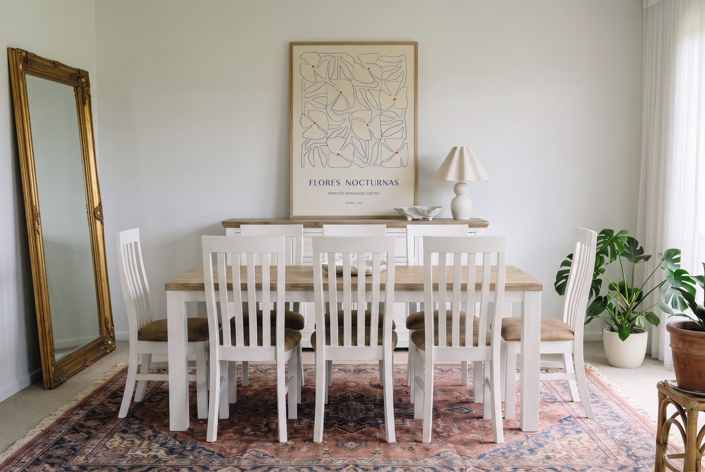Dining room with a wooden table, white chairs, vintage rug, gold floor mirror, and minimalist floral wall art.